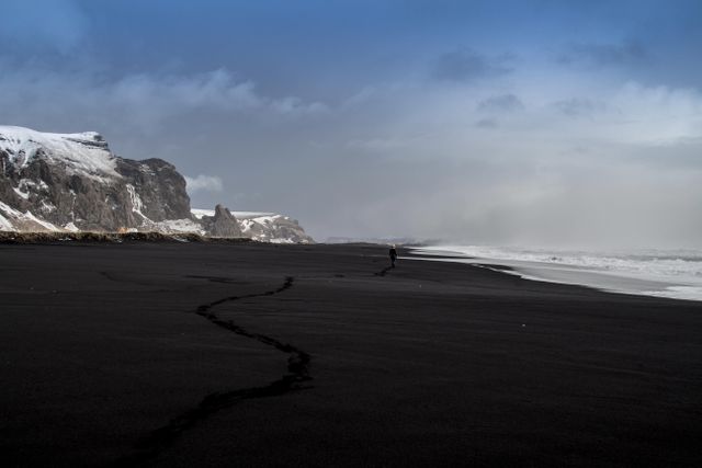 Person taking a solitary walk on a black sand beach with dramatic coastline in the background. Snow-covered cliffs rise to the left under moody sky. Ideal for travel blogs, adventure magazines, and websites promoting serene and remote locations. Can also be used in campaigns focusing on exploration, solitude, and nature's raw beauty.