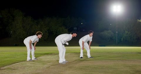 Night cricket fielders crouching under floodlights on pitch during tense match moment