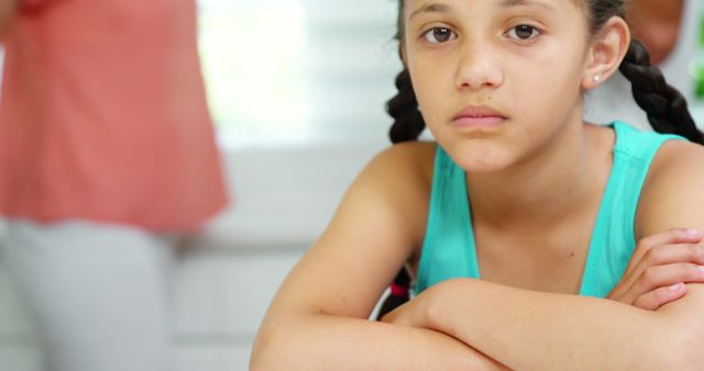 Sad girl with braids is leaning on a table indoors, displaying a serious expression. Image can be used to depict childhood emotions, family dynamics, or feelings of worry or contemplation. Suitable for articles, blogs, or advertisements about emotional wellness, child psychology, or family issues.