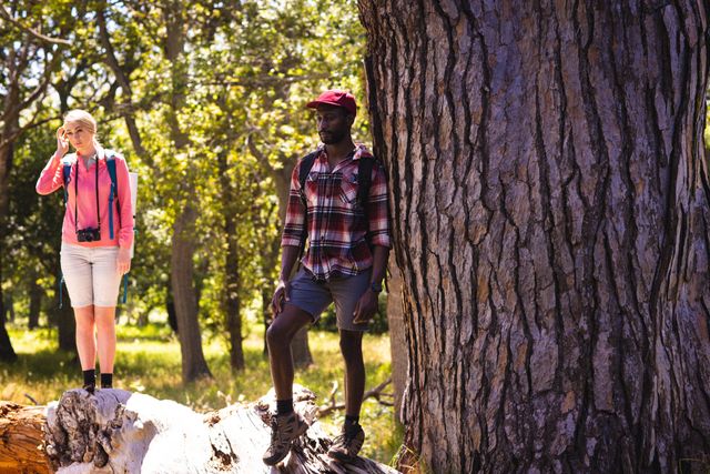 Diverse couple with backpacks leaning on tree in park on sunny day. Spending quality time, lifestyle and camping concept.