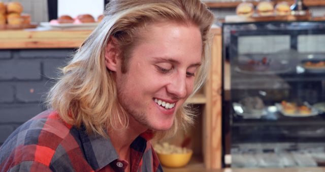 Young man with long blonde hair is smiling in a café wearing a plaid shirt. He looks happy and relaxed. Ideal for use in lifestyle blogs, café promotions, or casual fashion ads.
