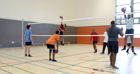 Diverse adult men playing competitive indoor volleyball spiking and blocking at gym