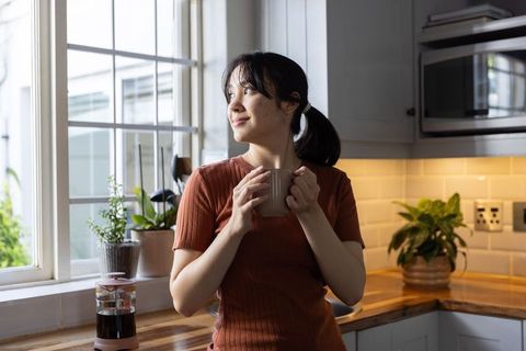 Asian woman enjoying morning coffee by sunlit kitchen window, relaxed domestic lifestyle