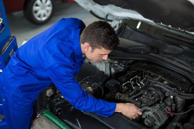 Young male mechanic in blue coveralls focused on repairing a car engine in a professional garage. Ideal for illustrating themes of automotive repair, maintenance services, professional workshops, and technical expertise. Can be used for automotive blogs, repair service advertisements, or maintenance tips.