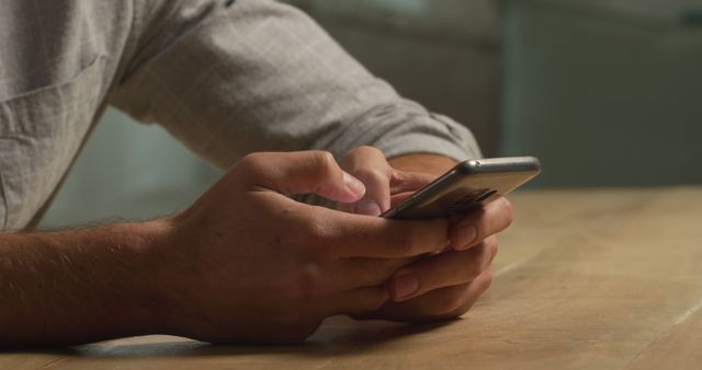 Close-up view of male hands holding and interacting with a smartphone while sitting at a table. Ideal for technology, communication, and remote work themes. Useful in articles and advertisements depicting everyday technology use, online communication, or mobile device interaction.