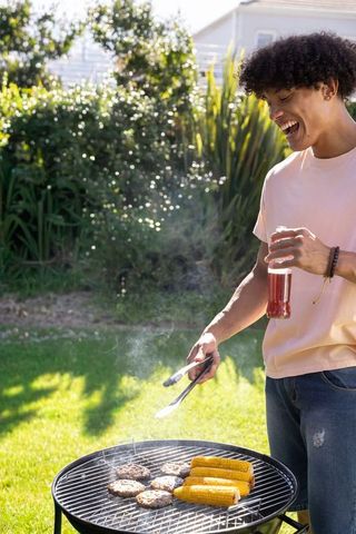 Young man grilling burgers and corn at sunny backyard barbecue, holding tongs and beverage