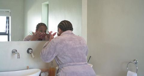 Mid-adult african american woman applying skincare at bathroom vanity in plush bathrobe