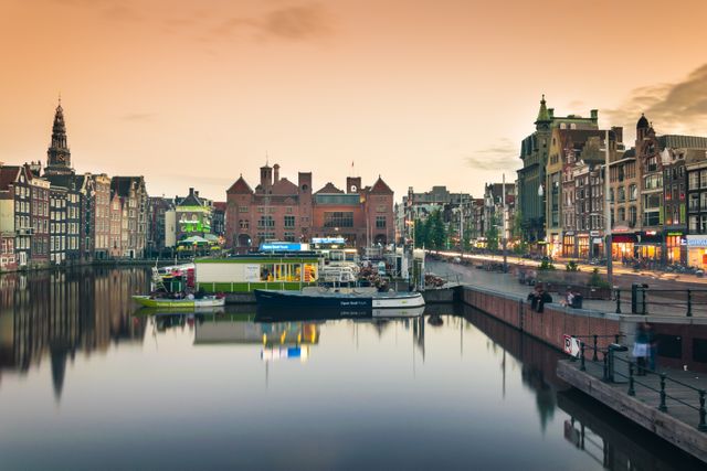 Scenic view of Amsterdam at sunset featuring a calm canal, historic buildings reflecting on the water, and boats docked. Perfect for promoting travel and tourism in the Netherlands, illustrating European urban landscapes, or enhancing a collection of scenic cityscape images.