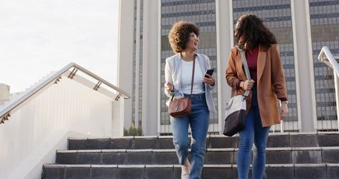 Walking and laughing on urban steps, friends chatting in smart casual with crossbody bags
