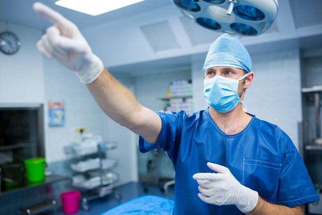 Surgeon in blue scrubs and mask pointing in an operating room. Ideal for use in healthcare, medical, and hospital-related content, illustrating surgical procedures, teamwork in healthcare, or medical professional environments.