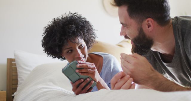 Image of happy diverse couple relaxing at home, lying on bed using smartphone and laughing. Happiness, communication, inclusivity, free time, togetherness and domestic life.