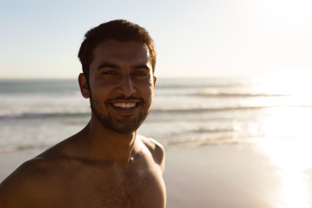 This image shows a young man smiling while standing on a sunny beach. The ocean waves and horizon are visible in the background, creating a serene and relaxing atmosphere. Ideal for use in travel brochures, vacation advertisements, lifestyle blogs, and wellness promotions.