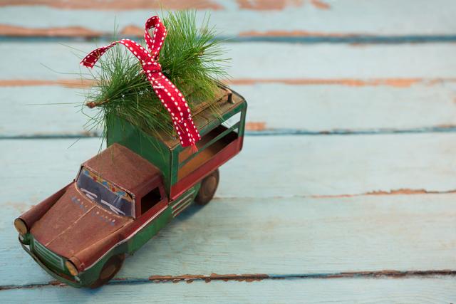 Toy truck carrying a small Christmas fir tree tied with a red ribbon on a rustic wooden plank. Perfect for holiday-themed designs, greeting cards, festive decorations, and seasonal marketing materials.