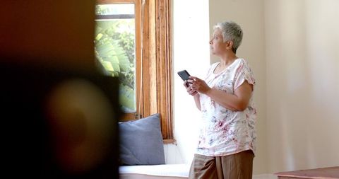 Thoughtful Senior Biracial Woman Using Smartphone by Window at Home