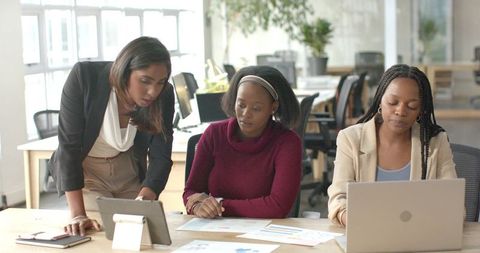 Diverse women collaborating in modern office reviewing tablet laptop and business charts