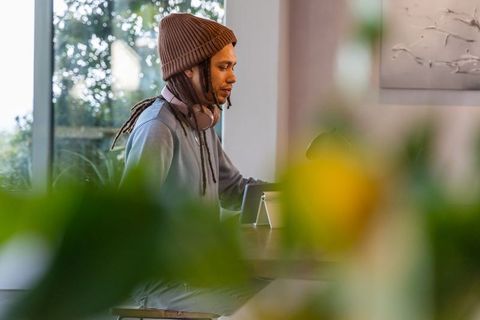 African american man using tablet and headphones at home workspace, cozy beanie and locs