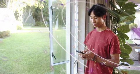 Asian teen using smartphone by open glass door overlooking backyard lawn, casual home lifestyle