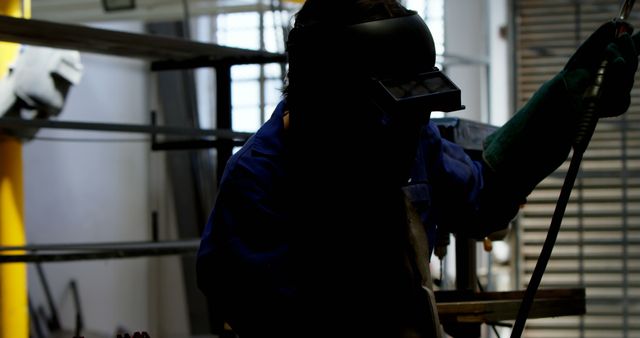 A silhouette of a worker wearing a welding mask is seen welding in a dimly lit industrial workshop. The scene highlights the craftsmanship and safety precautions taken during metalworking. Perfect for use in content related to manufacturing, industrial labor, engineering processes, safety standards in factories, and skilled trades.
