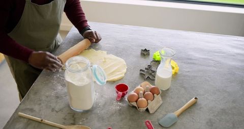 Mid adult male rolling cookie dough on gray countertop with flour eggs milk and baking tools