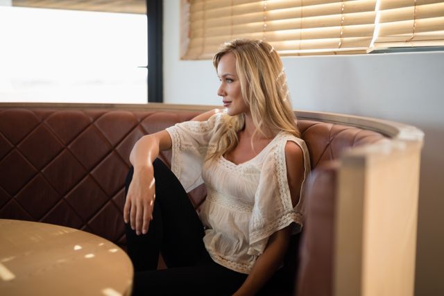 Woman sitting in a restaurant booth, looking thoughtful and relaxed. Ideal for use in lifestyle blogs, fashion articles, or advertisements promoting casual dining experiences. The natural light and serene expression convey a sense of peace and contemplation.