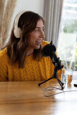 Cozy woman podcaster speaking into microphone wearing white headphones and mustard sweater