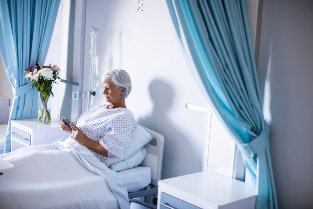 Senior woman in hospital bed using mobile phone, illustrating use of technology in healthcare. Ideal for healthcare, medical technology, patient care, and elderly lifestyle themes. Can be used in articles, advertisements, and educational materials about patient communication, hospital experiences, and recovery processes.