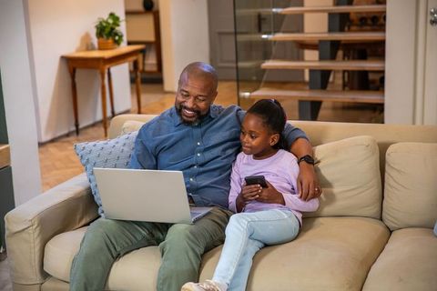 African american father and daughter relaxing on sofa using laptop and phone together