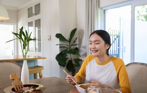 Asian woman enjoying healthy meal at bright home dining table with plants and chopsticks