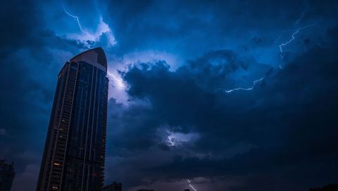 Modern skyscraper under lightning storm at night with dramatic blue clouds and lit windows