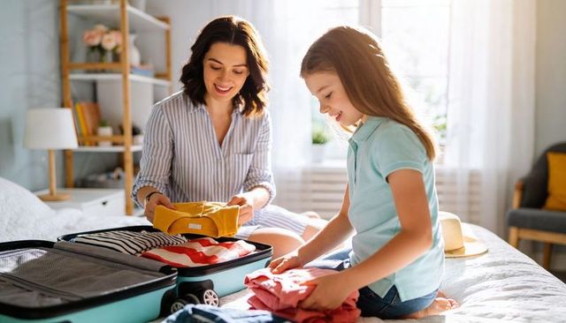Mother and daughter packing summer clothes into turquoise suitcase in bright bedroom
