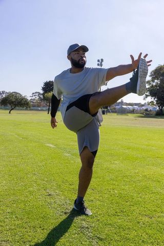 African american athlete performing high leg stretch on sunny park field for soccer training