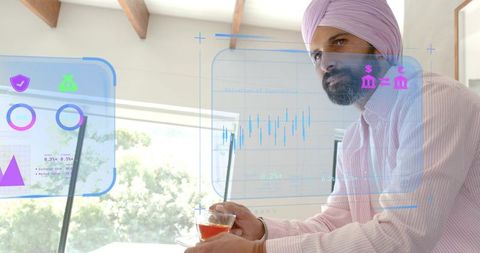 Man wearing pink turban holding tea reviewing holographic charts in home workspace