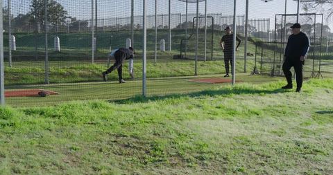 Mature men practicing baseball pitching in outdoor batting cages with coach observing
