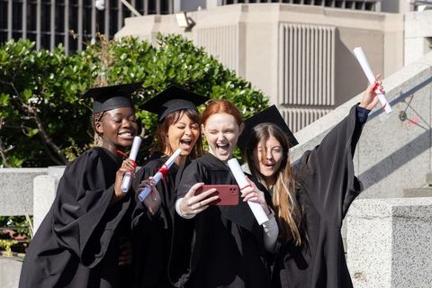 Diverse female graduates taking celebratory selfie on campus steps with diplomas in mortarboards