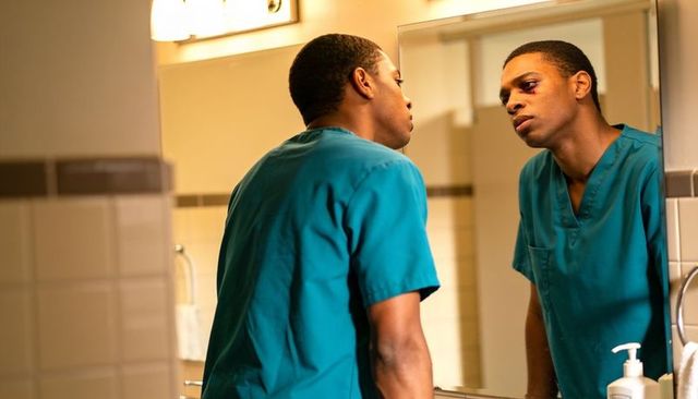 Tense medic inspecting bruised eye in bathroom mirror leaning on clinic sink wearing teal scrubs