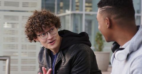 Curly haired student chatting with friend on urban campus bench, headphones visible
