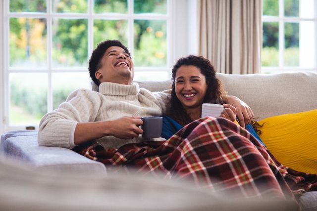 Cheerful biracial mid adult couple with coffee cups relaxing and chatting on sofa with blanket. Unaltered, love, togetherness, drink, winter, enjoyment and home concept.