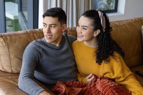 Young couple relaxing on leather sofa under blanket smiling and watching tv, cozy moment