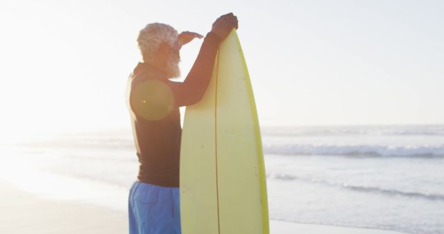 Older man with surfboard standing on beach looking at ocean horizon. Perfect for promoting active aging, healthy lifestyles, water sports programs for seniors, and advertisements featuring fitness and seaside leisure.