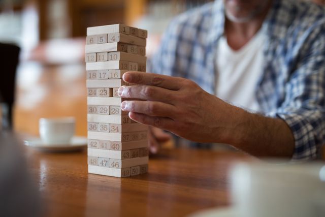 Man's hand carefully removing a block from a Jenga tower in a bar. Ideal for illustrating concepts of leisure activities, strategy games, concentration, and casual social settings. Suitable for use in articles, blogs, and advertisements related to social gatherings, bars, and indoor games.