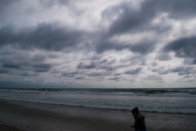 Person walking along stormy beach under cloudy sky creates a moody atmosphere with dark clouds and ocean waves. This image is perfect for concepts related to solitude, contemplation, nature, and weather. It can be used for blog post images, social media graphics, and thematic websites focusing on nature, emotions, or coastal environments.