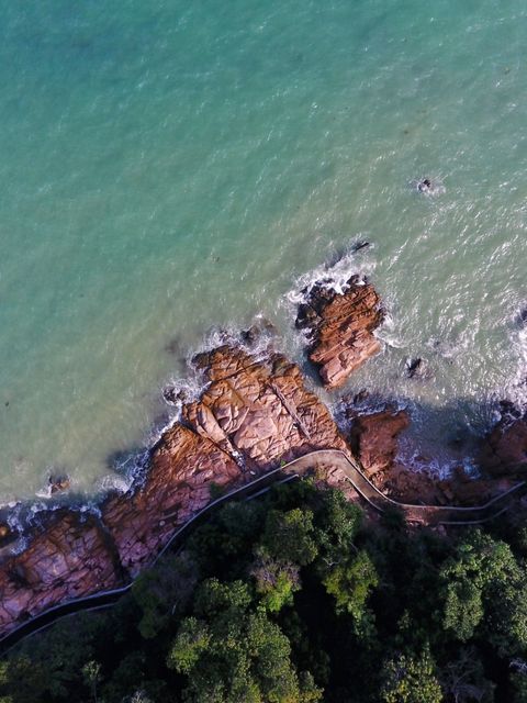 Aerial view capturing rocky coastline meeting turquoise ocean waters, bordered by lush greenery. Ideal for use in travel brochures, nature documentaries, and scenic calendars. Emphasizes the natural beauty and tranquil environment of coastal regions.