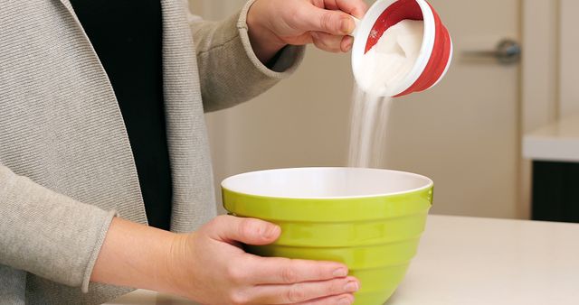 This close-up scene captures a person pouring sugar from a measuring cup into a green mixing bowl. It conveys the beginning of a baking or cooking process, emphasizing preparation and the use of ingredients. This image is suitable for culinary blogs, recipe websites, food-related advertisements, cooking classes, and educational materials focusing on kitchen activities.