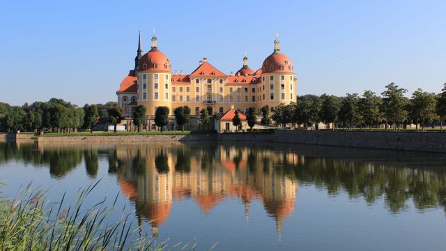 Baroque Moritzburg Castle with distinctive red domes situated in Germany, reflected perfectly in the calm waters of the surrounding lake on a sunny day. Ideal for travel brochures, tourism promotion, historical architecture studies, scenic calendars, and educational content on European cultural heritage.