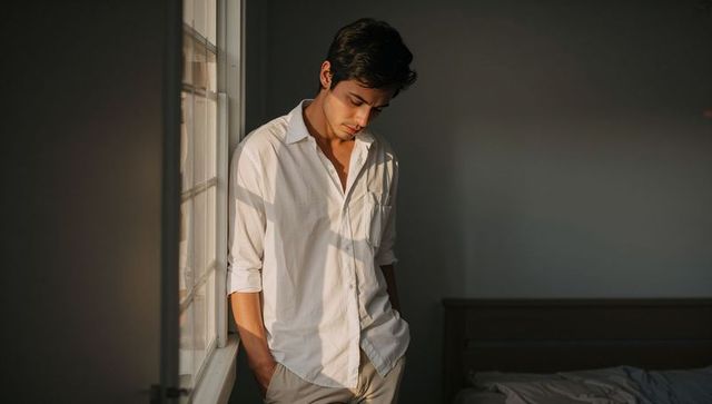 Contemplative young man by sunlit multi-pane window in minimalist bedroom interior