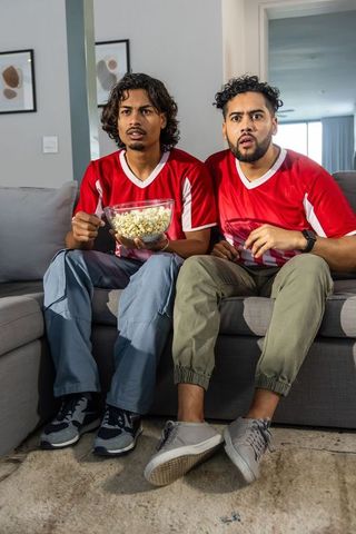 Diverse friends watching soccer match on couch holding popcorn reacting with surprise