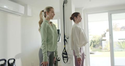 Two women standing for home workout with trx and kettlebells in sunlit minimalist gym