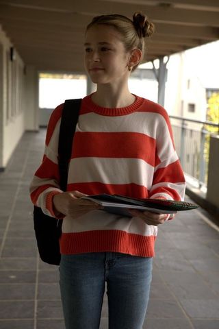 Schoolgirl holding notebooks and backpack standing in school corridor wearing red striped sweater