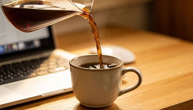 Pouring morning coffee from glass carafe into ceramic mug on wooden desk with laptop