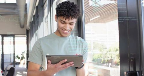 Young man smiling while using tablet with code reflections in sunlit modern office